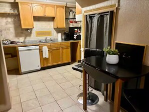 Una cocina compacta con armarios de madera, lavavajillas, cafetera y una pequeña mesa de comedor con planta en maceta.