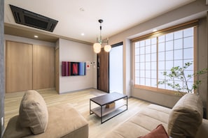 A modern living room with a sofa, a coffee table, a large window with a view, and a door leading to another room. - Shinjuku 6-chome City Stay 1F (Shinjuku)