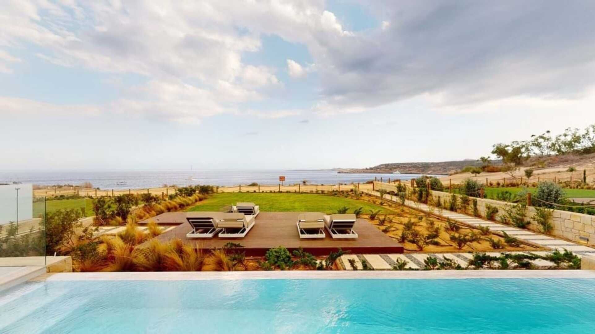 A poolside view with a clear sky, a beach in the distance, and a well-maintained lawn with lounge chairs.