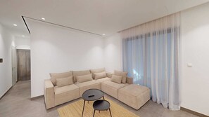 Un salon moderne avec un canapé d’angle, une petite table et une grande fenêtre ornée de rideaux légers.