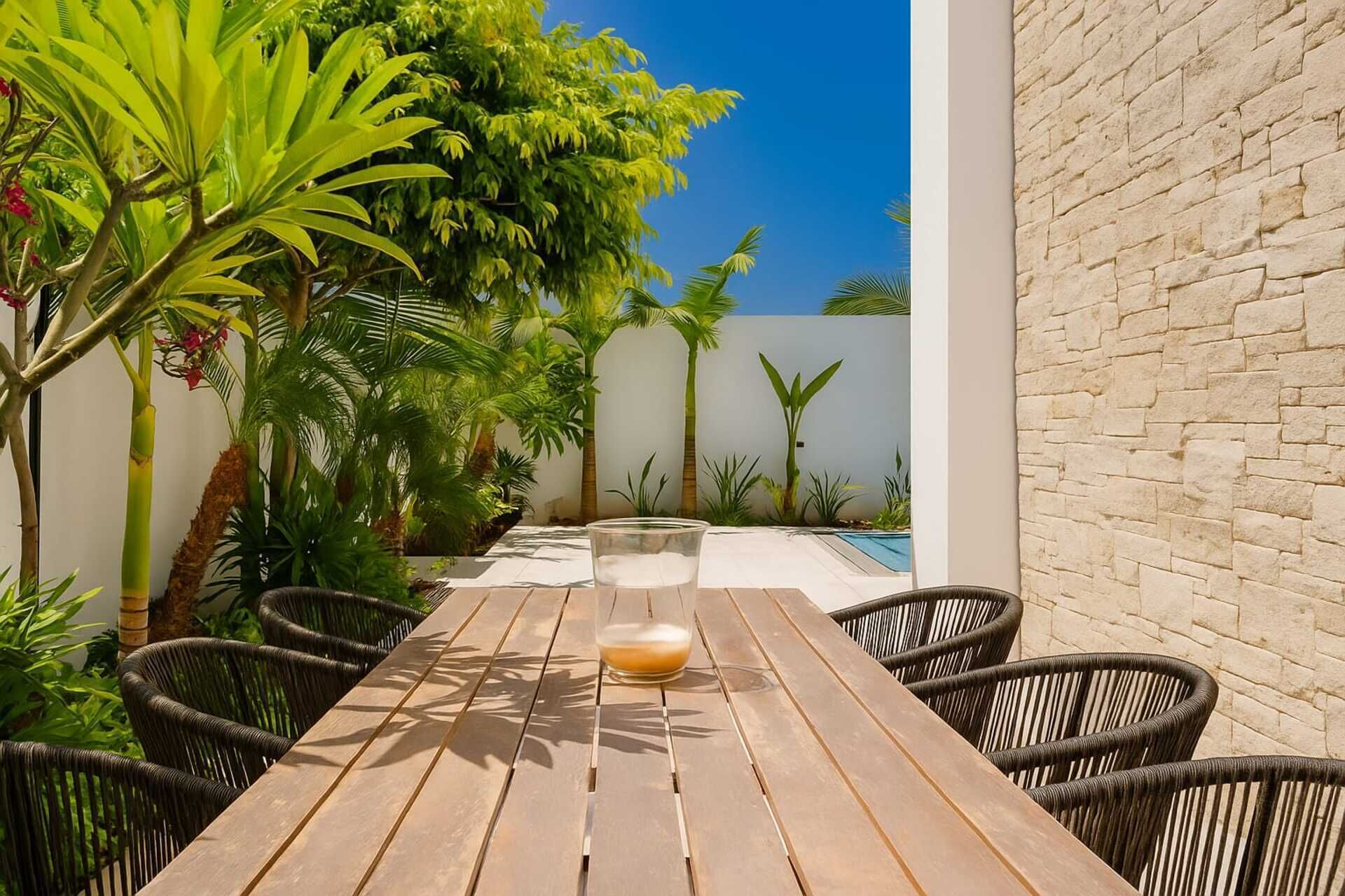 An outdoor patio with a wooden table and wicker chairs, surrounded by lush greenery and a stone wall.