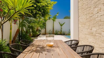 An outdoor patio with a wooden table and wicker chairs, surrounded by lush greenery and a stone wall.