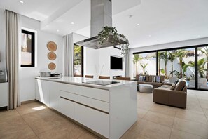 A modern kitchen with a central island, a dining area with a sofa, and a large window with a view of palm trees.