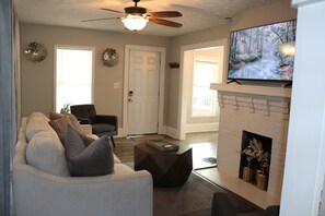 A living room with a fireplace, a television mounted above it, a ceiling fan, and a door leading to another room.