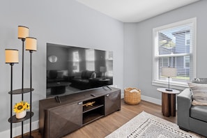 A modern living room with a flat-screen TV, dark wood furniture, a grey sofa, and a large window with blinds. - 7 Minutes to Campus! (Gainesville)
