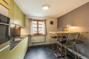 A compact kitchen with a dining area, a window with curtains, a radiator, and a clock on the wall. - Luxury Alpine Retreat|Zermatt 1068 (Wallis)