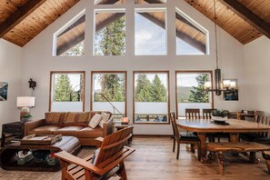 A spacious living room with a high ceiling, large windows, a brown leather sofa, a dining table with chairs, and a wooden floor. - Whispering Pines Sanctuary near Payette River & Hot Springs (Garden Valley)
