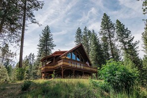 Eine Holzhütte mit einer Holzterrasse und großen Fenstern, umgeben von hohen Kiefern.
