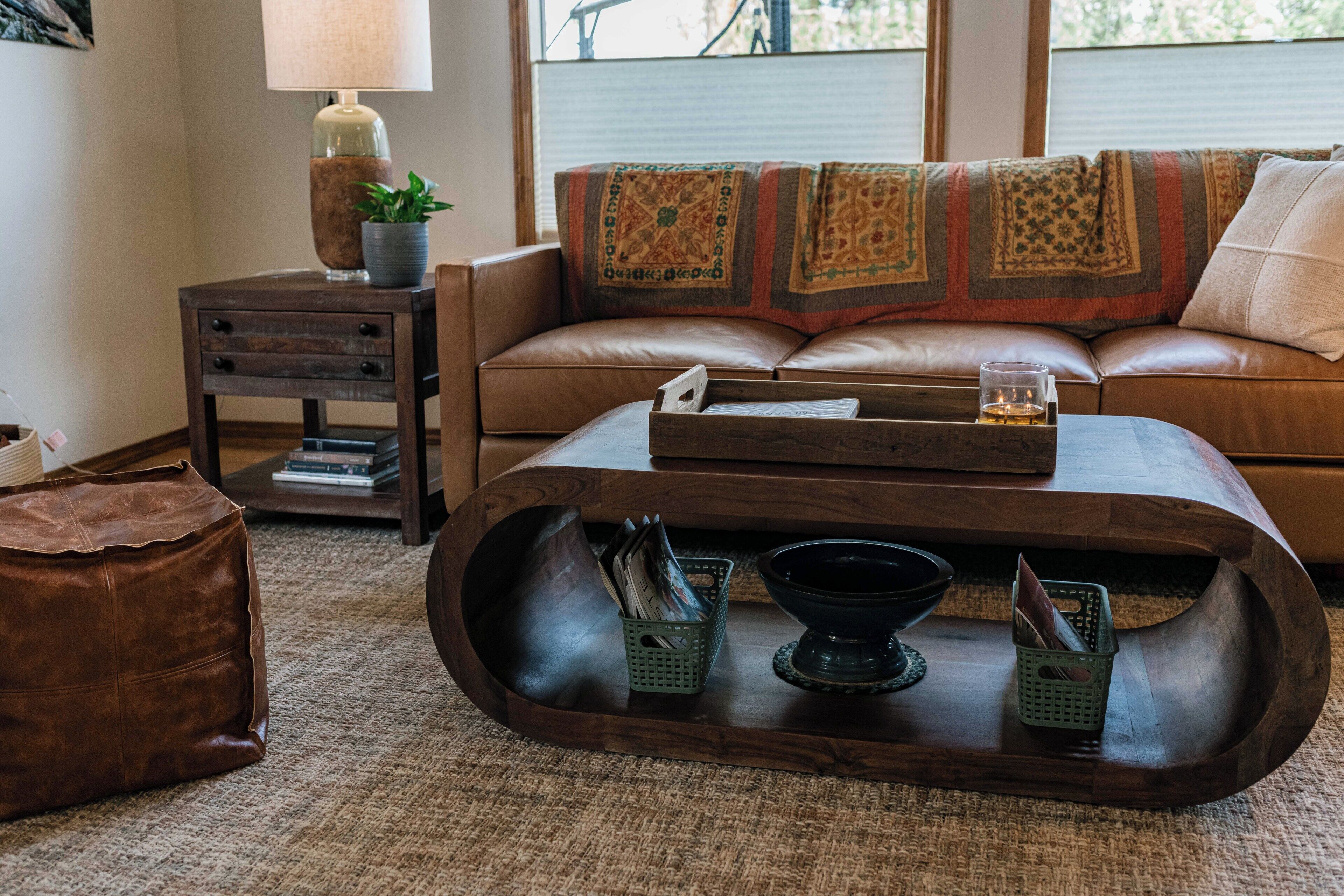 A cozy living room with a leather sofa, a wooden coffee table with magazines, a lamp, and a window with blinds.