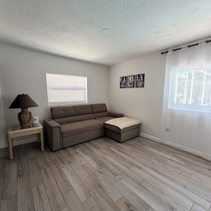 Ein Zimmer mit einem Ecksofa, einer Lampe, einem kleinen Tisch und einem Fenster mit Jalousien.