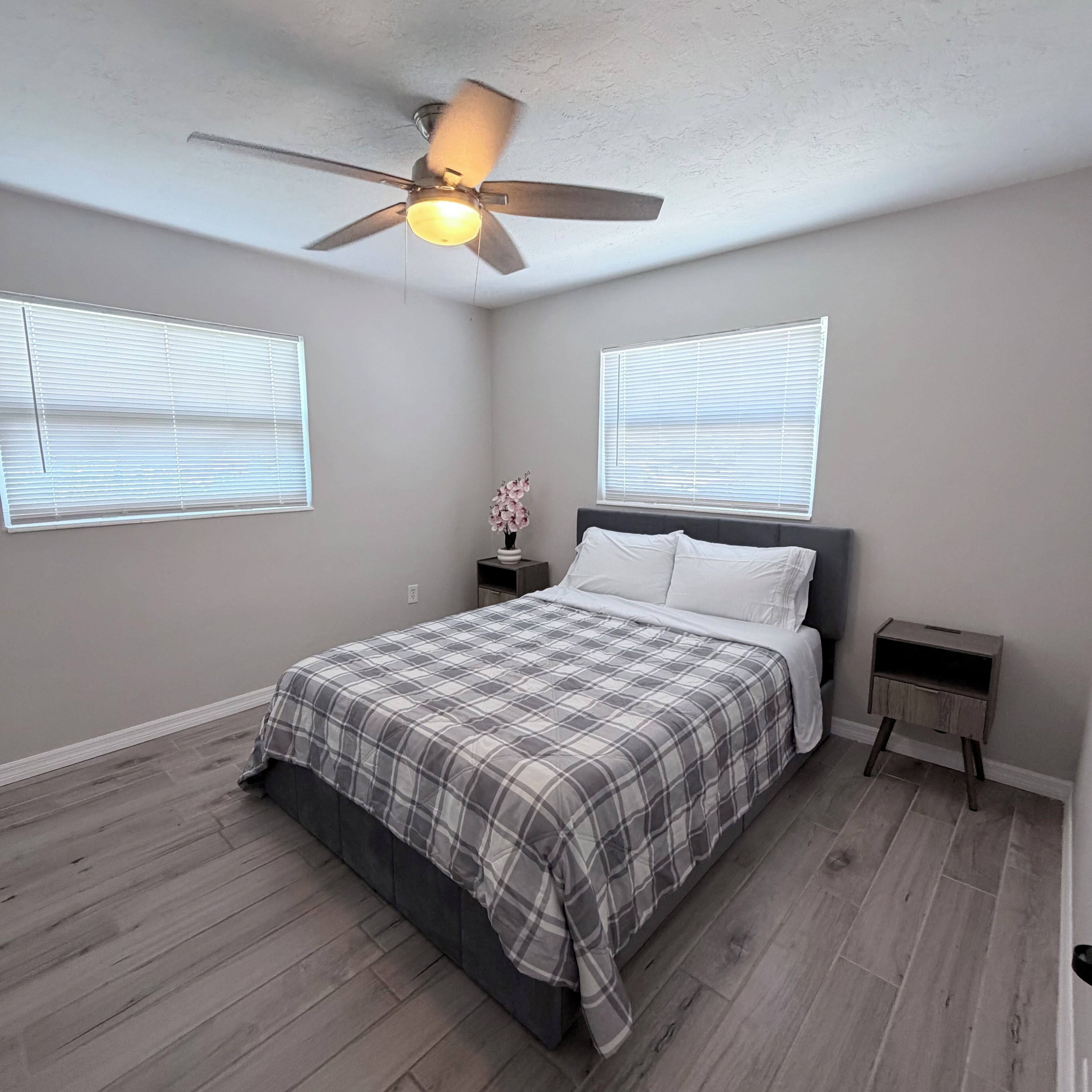 Un dormitorio con cama, mesitas de noche, ventilador de techo y dos ventanas con persianas.