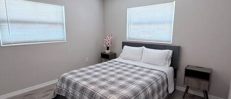 Un dormitorio con cama, mesitas de noche, ventilador de techo y dos ventanas con persianas.