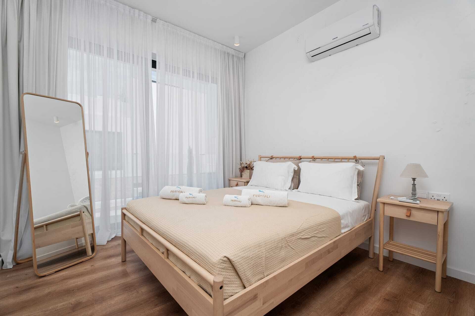 Ein schlicht eingerichtetes Schlafzimmer mit einem hölzernen Bett, weißen Bettwäsche, einem Nachttisch mit Lampe und einer Wandaircondition.