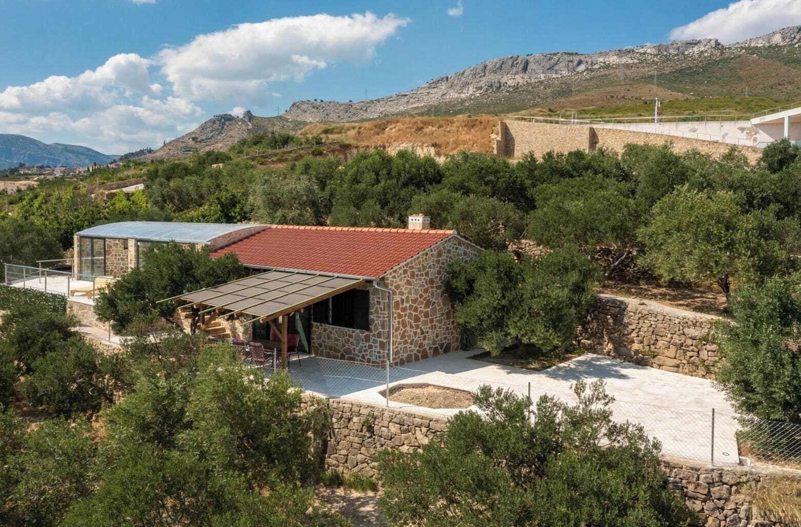 A stone house with a red-tiled roof, surrounded by trees and a mountainous backdrop.