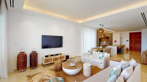 Ein modernes Wohnzimmer mit Flachbildfernseher, weißen Sofas, einem Couchtisch und einem Essbereich mit Küche im Hintergrund.