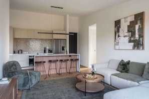 Ein modernes Wohnzimmer mit einem grauen Sofa, einem runden Couchtisch mit Früchten und einer Küche im Hintergrund.