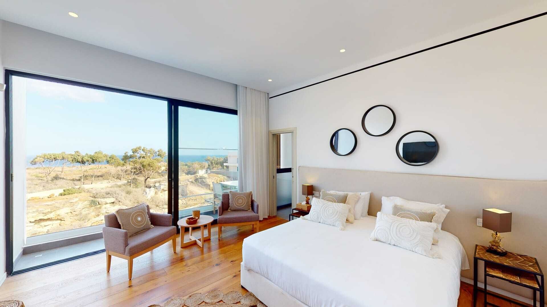 A modern bedroom with a large bed, a seating area, and a view of the landscape.