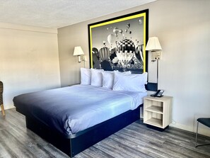 A modern hotel room with a large bed, a headboard with a black and yellow frame, two bedside lamps, a small wooden nightstand, and a wall art depicting hot air balloons. - Super 8 Downtown_1 King Bed (Albuquerque)