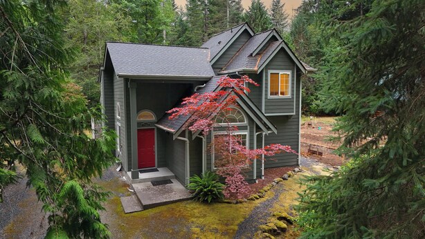 Una casa petita de dues plantes amb una porta vermella i un petit porxo, envoltada per un bosc dens.