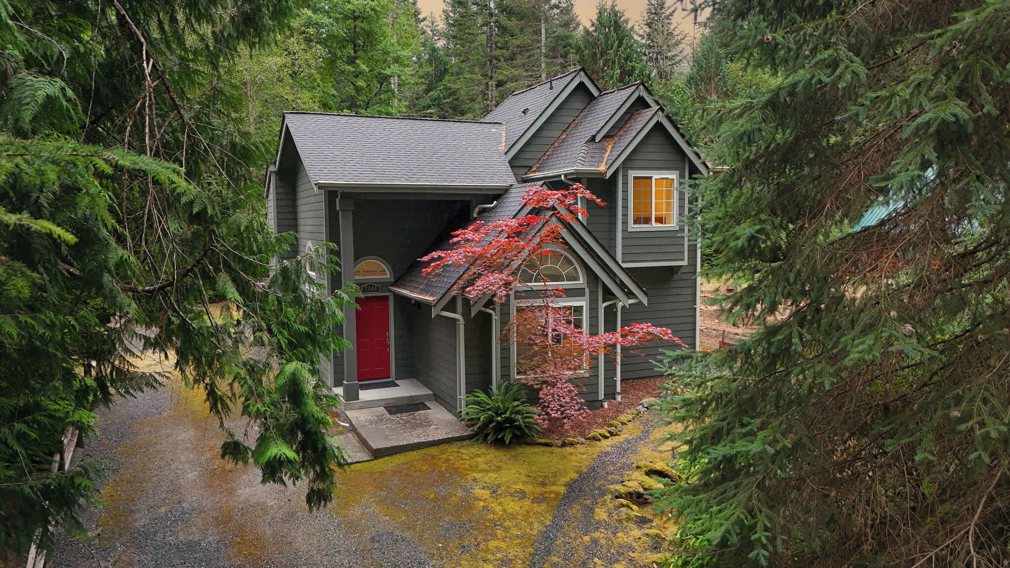 A two-story house with a red door, surrounded by dense forest.