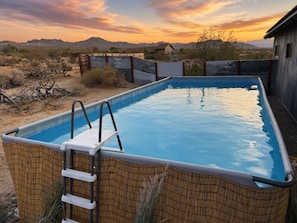 Una piscina rectangular con escalera, rodeada por una cerca de madera, ubicada en un paisaje desértico con montañas al fondo.