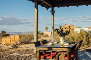 An outdoor dining area with a table set for two, surrounded by chairs, a potted plant, and a view of the desert landscape. - Park-edge hot tub retreat · dogs welcome (Joshua Tree)
