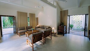 A spacious living room with a sofa set, a coffee table, and a wooden chair. There is a sliding glass door leading to a balcony with a view of greenery.
