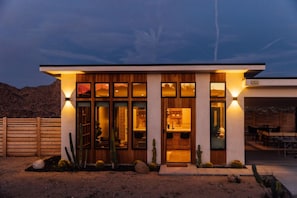 A modern house with a wooden facade, large windows, and a well-lit entrance. - Casa Mariposa| 5 Minutes to Park Entrance| Hot Tub (Joshua Tree)