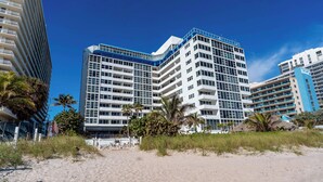Ein Strandhotel mit mehreren Etagen, ein Sandstrand im Vordergrund und Palmen.