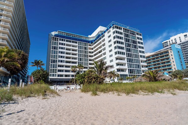 Un hotel frente a la playa con varios pisos, una playa arenosa al frente y palmeras.