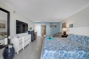 A hotel room with a large bed, a TV mounted on the wall, a minibar, and a view of the beach.