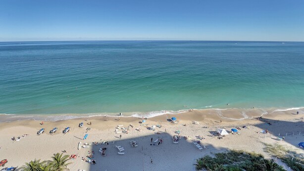 Ein Strand mit blauem Himmel, ruhigem Wasser und einem sandigen Ufer mit Liegestühlen und Sonnenschirmen.