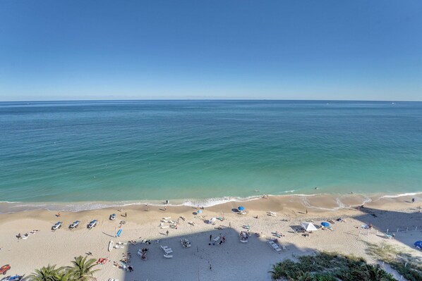 Ein Strand mit blauem Himmel, ruhigem Wasser und einem sandigen Ufer mit Liegestühlen und Sonnenschirmen.