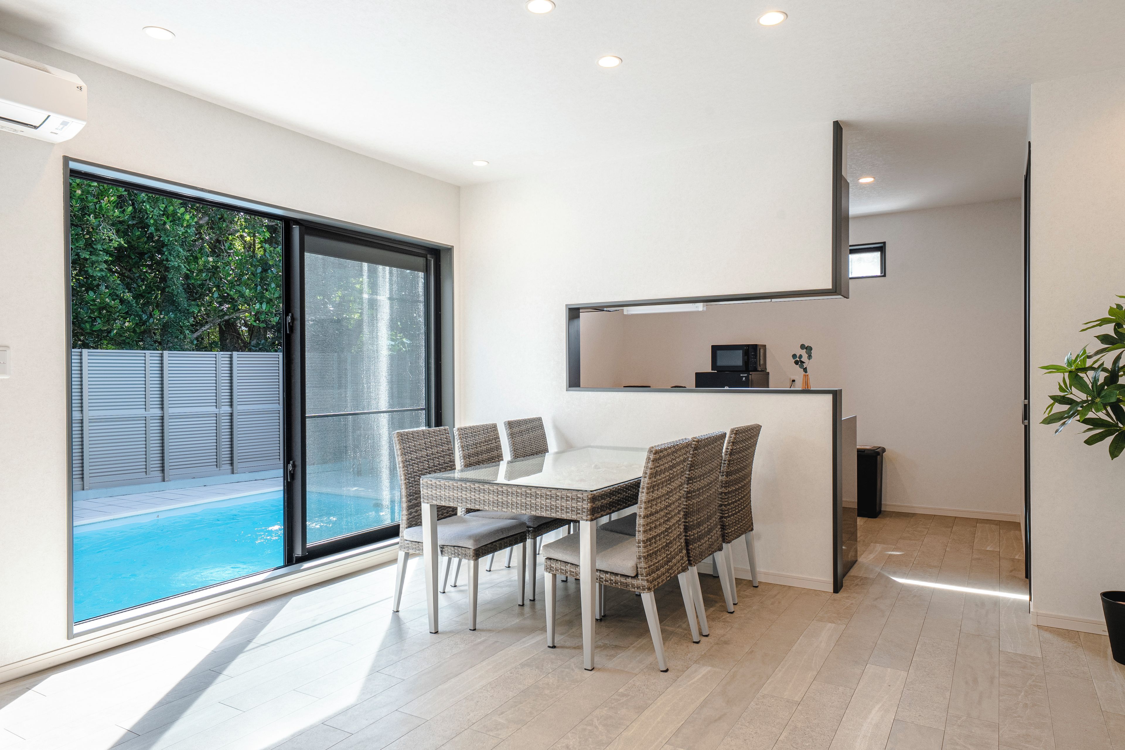 A modern dining area with a glass table and wicker chairs, a small kitchen area with a microwave, and a view of a swimming pool outside.