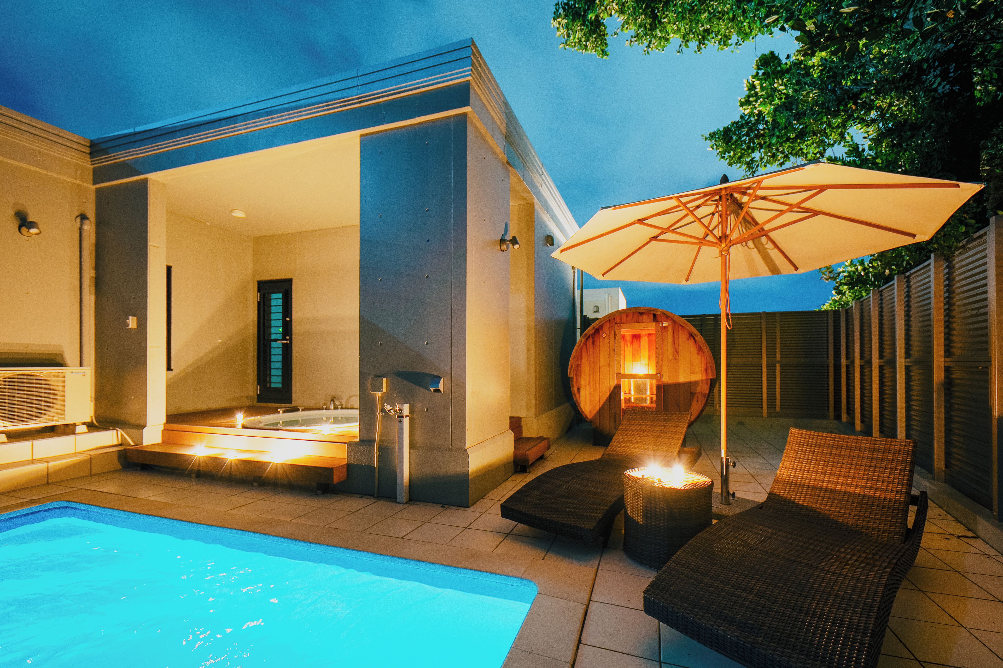 Un área de piscina moderna al aire libre con un jacuzzi, sillas de descanso y una sombrilla grande.