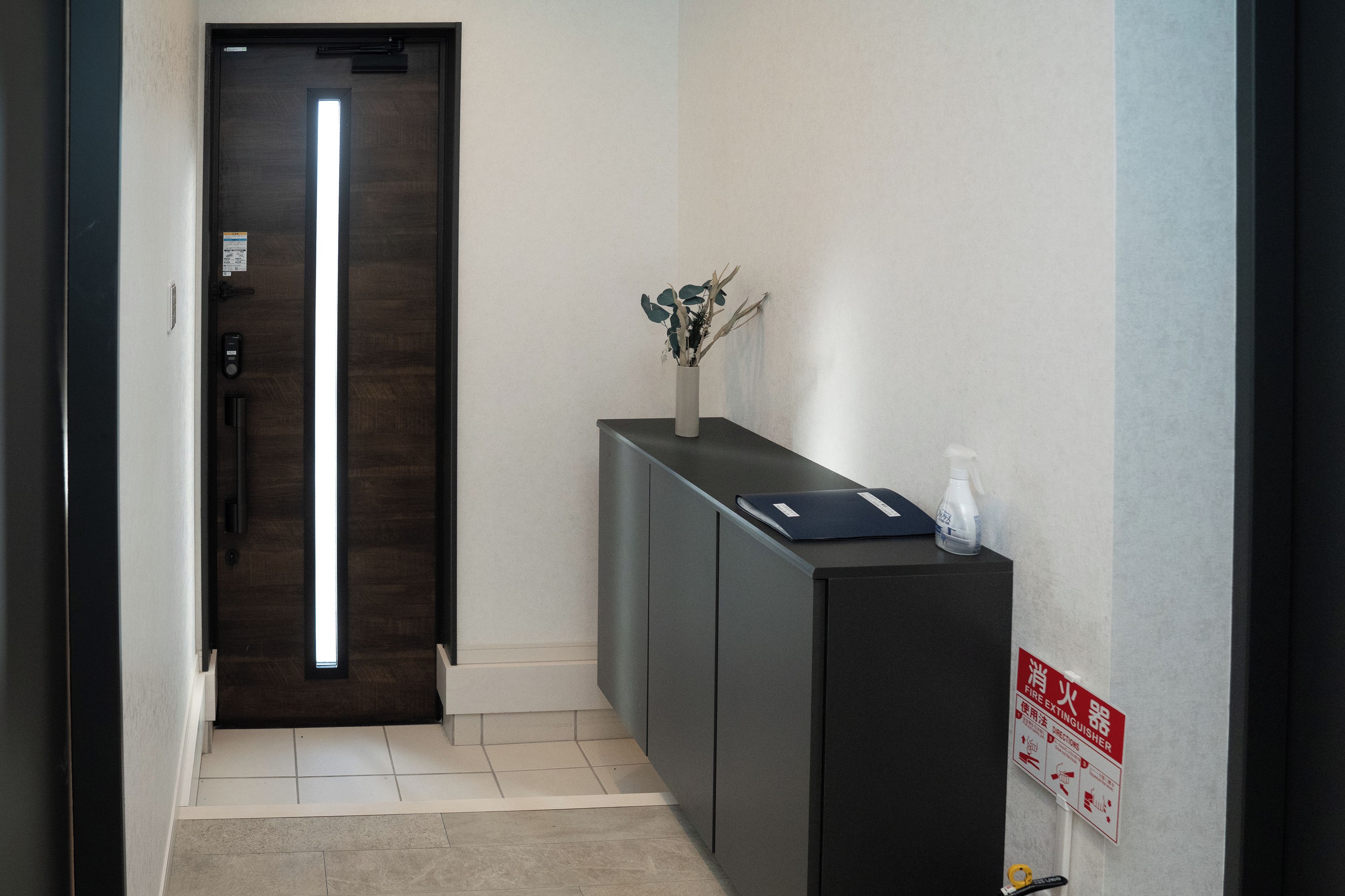 Un couloir avec une porte en bois sombre, un comptoir d’accueil orné d’un vase de fleurs et un panneau indiquant l’emplacement d’un extincteur sur le mur.