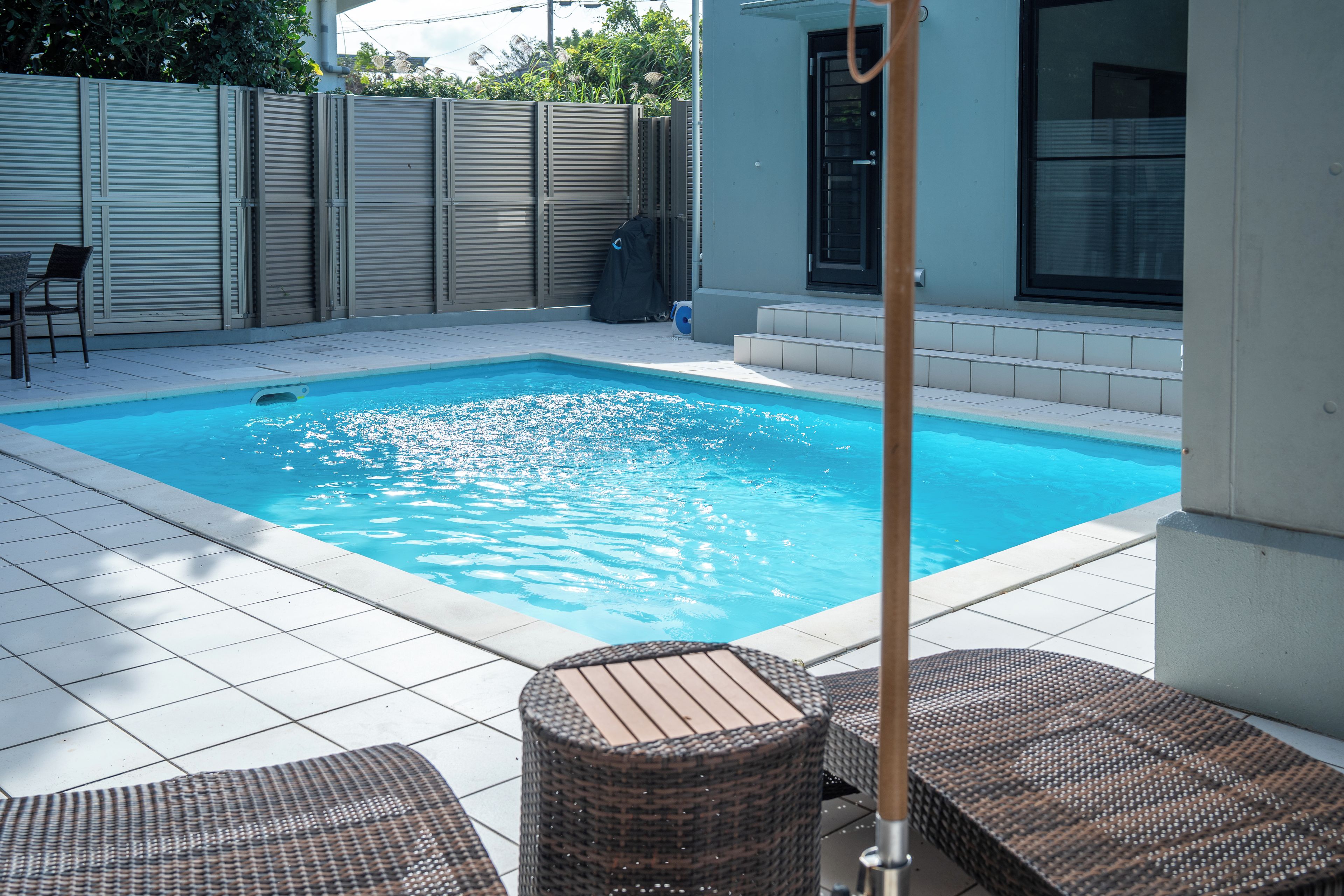 Piscina rectangular con solárium de baldosas, rodeada por una cerca baja y un porche cubierto con muebles de mimbre.