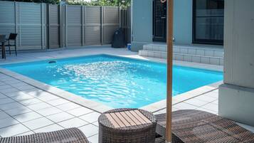 Piscina rectangular con solárium de baldosas, rodeada por una cerca baja y un porche cubierto con muebles de mimbre.