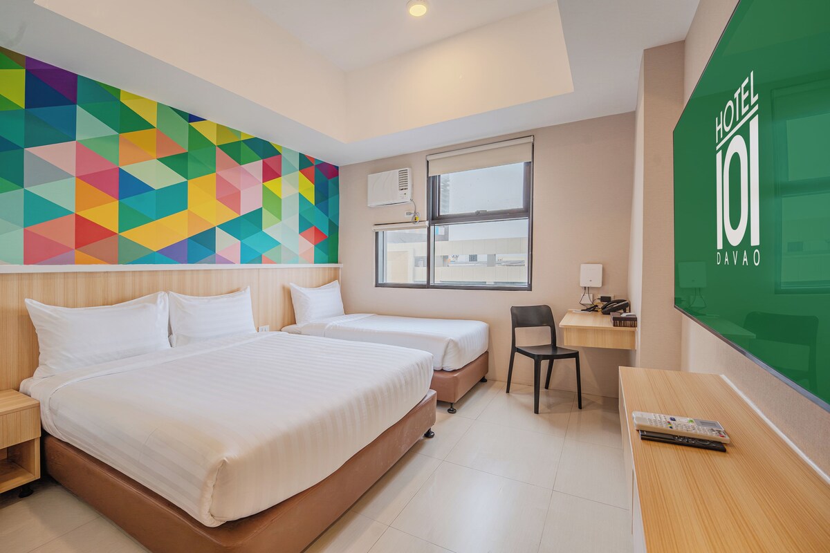 A hotel room with two beds, a desk, a chair, a colorful geometric wall, and a window.