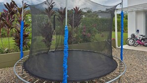 Un trampolín con red de seguridad, rodeado de plantas y una bicicleta al fondo.