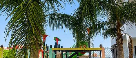 A playground with slides and climbing structures, surrounded by palm trees and a fence.
