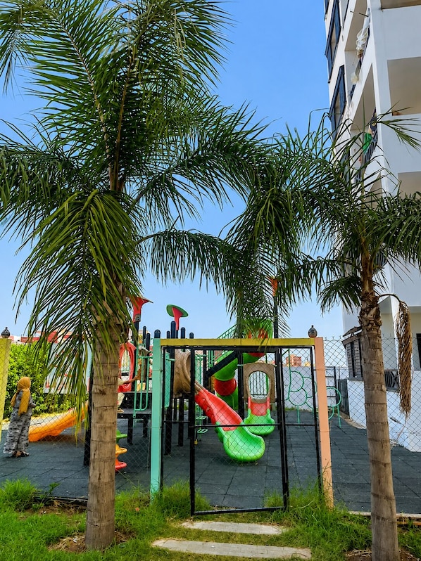 A playground with slides and climbing structures, surrounded by palm trees and a fence.