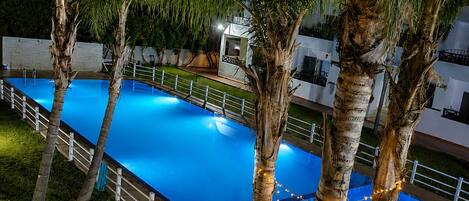 A swimming pool surrounded by palm trees and a white fence, with a small table and chairs nearby.