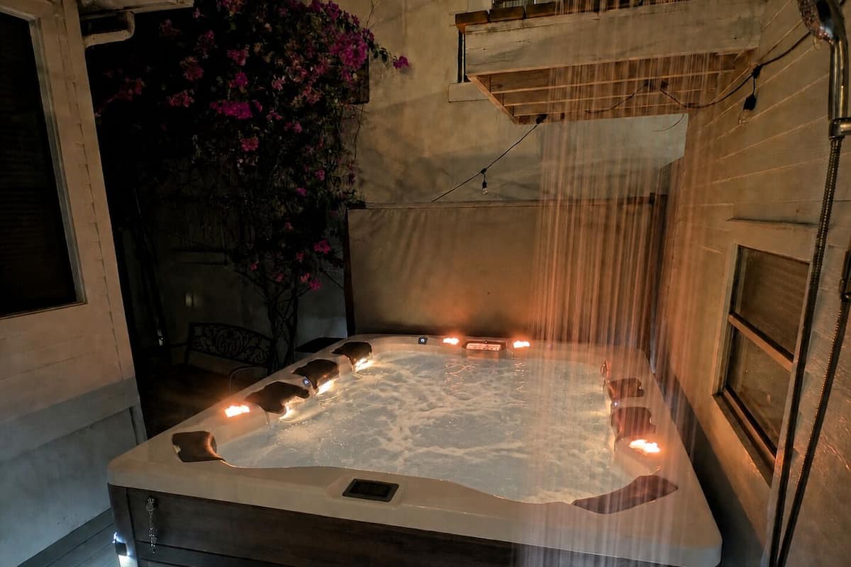 Ein Whirlpool mit einer Holzbank, einer an der Wand befestigten Duschbrause und einem Topf mit rosa Blüten.