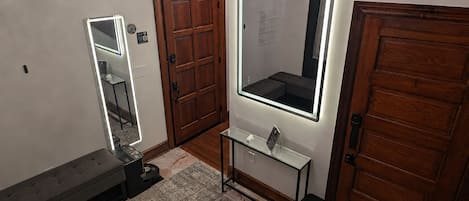 A bathroom with a wooden door, a mirror with integrated lighting, a bench, and a small table with a sink.