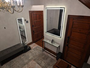 A bathroom with a wooden door, a mirror with integrated lighting, a bench, and a small table with a sink.