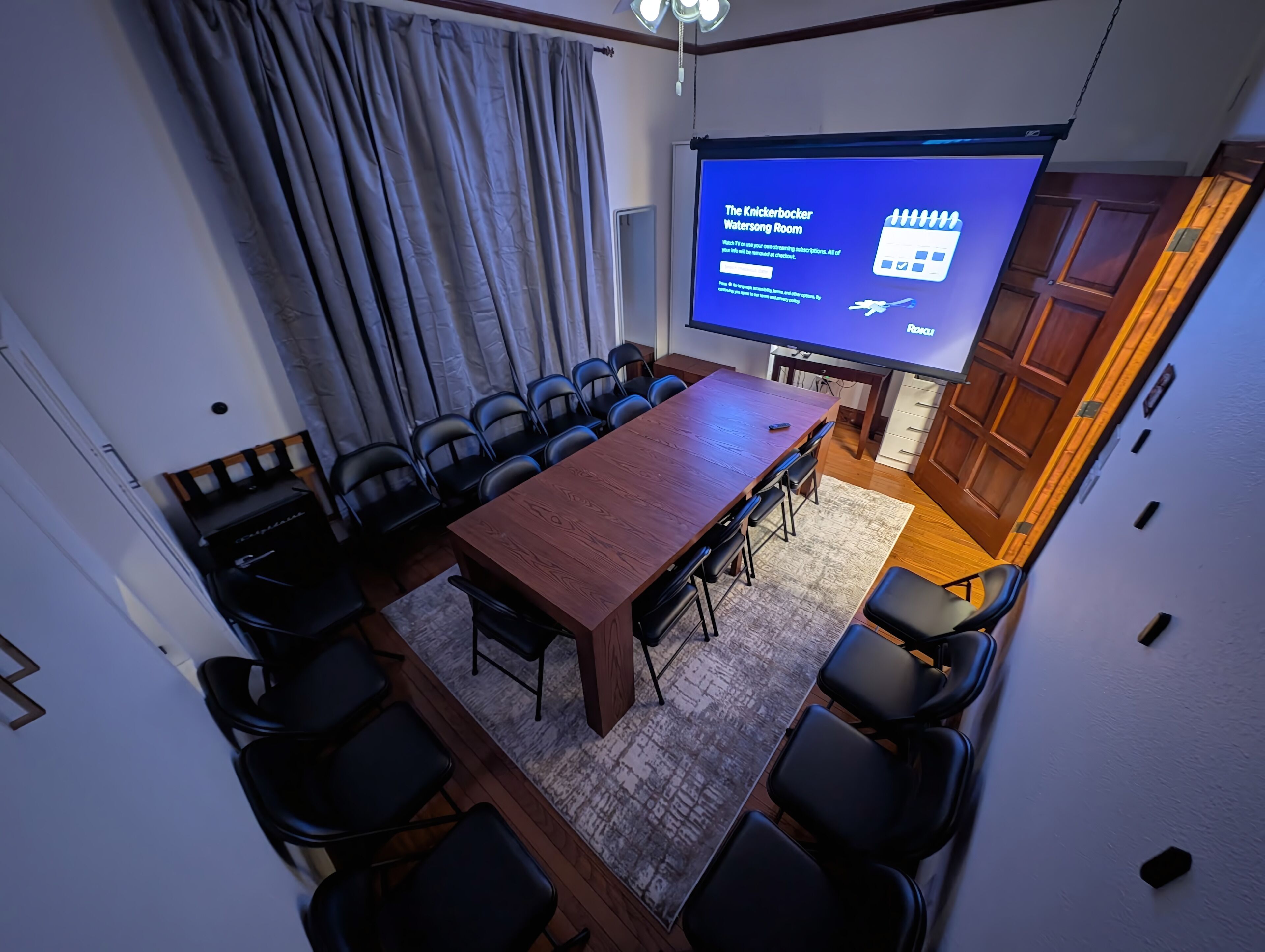 A conference room with a large screen displaying 'The Knickerbocker Watersong Room' text, a wooden table, and black chairs arranged around it.