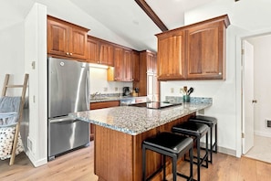 A kitchen with wooden cabinets, a stainless steel refrigerator, granite countertops, and a bar stool area. - Four Seasons Retreat | Minutes to Skiing and Downtown (Ketchum)