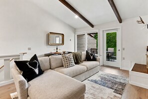 A modern living room with a sectional sofa, a coffee table, and a rug. There is a dining area visible through an open door. - Four Seasons Retreat | Minutes to Skiing and Downtown (Ketchum)