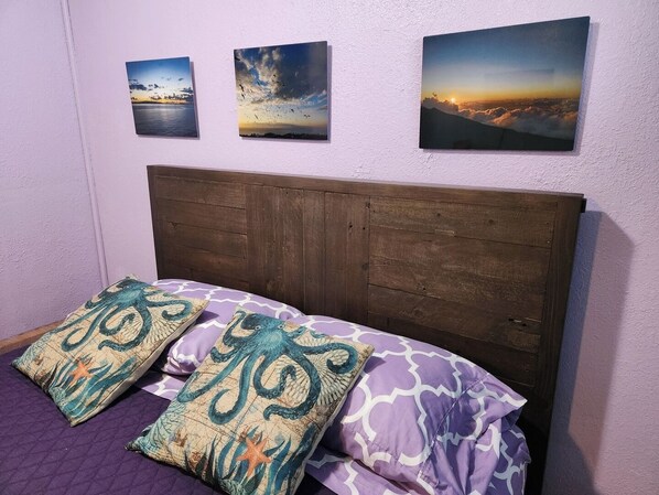Ein Schlafzimmer mit einem hölzernen Kopfende, einer violetten Tagesdecke und drei Landschaftsbildern an der Wand.